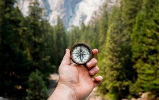 A hand holds up a compass in the middle of the woods